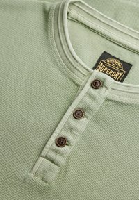 Maglietta henley a maniche lunghe, verde chiaro e texturizzata, con placca a bottoni e bottoni marroni. L'etichetta riporta "Superdry" su sfondo nero.