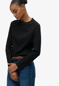 Zwarte long sleeve top met een ronde hals, voorzien van een klein logo op de borst. Gepaard met blauwe jeans en een bruine gevlochten riem.