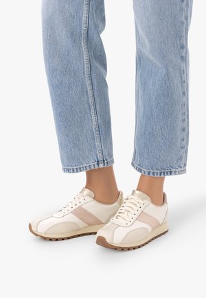 Stopy w białych i beżowych sneakersach w połączeniu z jasnoniebieskimi skróconymi dżinsami na białym tle.
