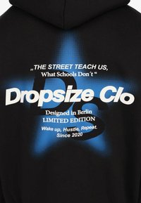 Zwarte hoodie met een blauwe stergrafiek en witte tekst: "De straat leert ons wat scholen niet doen," Dropsize Clo, ontworpen in Berlijn, limited edition sinds 2020.