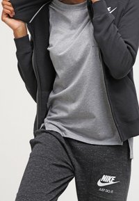 Černá zipová bunda, šedé triko s kulatým výstřihem a tmavě šedé tepláky s bílým logem Nike. Měkká látka, uvolněný střih a viditelné švy.