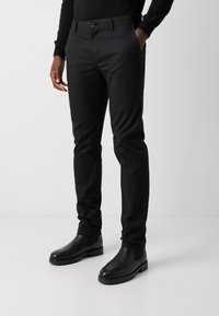 Zwarte stretchbroek met een slim fit, voorzien van een klassieke knoopsluiting en zijzakken. G draagt zwarte leren chelsea boots.