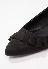 Chaussure plate noire à bout pointu avec un détail de volants pliés sur le devant et une texture douce ressemblant à du daim.