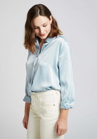 Blouse en satin bleu clair avec un col pointu, fermeture à boutons et manches longues retroussées. Présente un motif à pois subtil et une texture lisse.