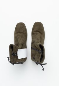 Botas de ante verde oliva hasta el tobillo con punta cuadrada, cierre de cremallera lateral y detalles de lazo en la abertura. Etiquetas visibles en la bota izquierda.