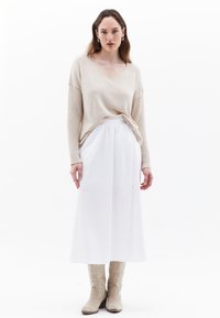 Beige gestrickter Pullover mit V-Ausschnitt, kombiniert mit einem fließenden weißen Rock. Die Schuhe bestehen aus leichten, strukturierten kniehohen Stiefeln.