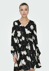 Robe noire avec un motif floral abstrait blanc, décolleté en V, manches longues, taille ceinturée et jupe fluide. Tissu lisse avec un léger éclat.