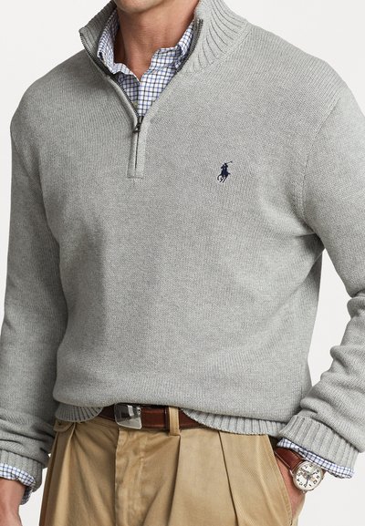 Hellgrauer Strickpullover mit einem Viertelreißverschlusskragen, gerippten Bündchen und einem gestickten Logo. Getragen über einem karierten Hemd und khakifarbenen Hosen.