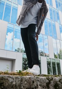 Grå pufferjakke, hvid t-shirt, sorte sweatpants med sideliste, og hvide sneakers. Står på en sten med grønne planter i baggrunden.