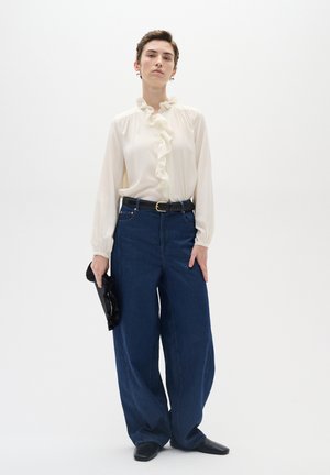 Cremefarbene Bluse mit Rüschenelementen an der Vorderseite, leichtes Material; dunkelblaue weit geschnittene Jeans; schwarze Clutch; kurze Haare; stehend vor einem weißen Hintergrund.