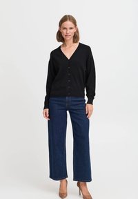 Cardigan nero a maglia con scollatura a V e bottoni frontali, abbinato a jeans in denim scuri a gamba larga e tacchi beige. Tessuto liscio e vestibilità comoda.
