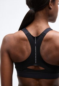 Πίσω όψη ενός ατόμου που φοράει μαύρο αθλητικό σουτιέν racerback με το κείμενο "SOUND MIND SOUND BODY" κατά μήκος της σπονδυλικής γραμμής.