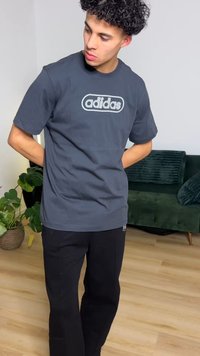 Tumši pelēka kokvilnas T-krekls ar centrālu baltu Adidas logotipu. Redzamas melnas bikses, iestādītas gaismas koka grīdas interjerā.