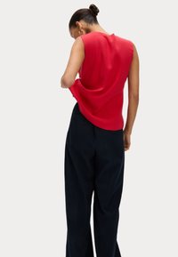 Rotes ärmelloses Oberteil aus weichem Material mit lockerer Passform. Kombiniert mit dunklen Nadelstreifenhosen. Dezenter Glanz auf dem Oberteil.
