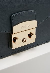 Grå läderhandväska med texturerad yta, utrustad med en guldtonad låsning med dubbla runda knappar och ingraverat varumärkesnamn på locket.