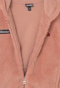 Polarowa kurtka zapinana na zamek w jasnoróżowym kolorze o miękkiej fakturze, z logo na czarnej naszywce i kolorowym akcentem przy zamku.