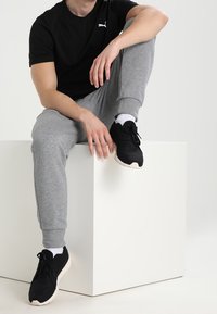 Svart Puma-t-shirt, grå joggingbyxor, svarta sportskor med vita sulor, sitter på en vit kub. Fokusera på avslappnad, casual sportklädsel.