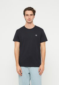 Čierne bavlnené tričko s krátkymi rukávmi, okrúhlym výstrihom a malým logom na hrudi. Hladká textúra, štandardný strih, v kombinácii so svetlomodrými džínsami.