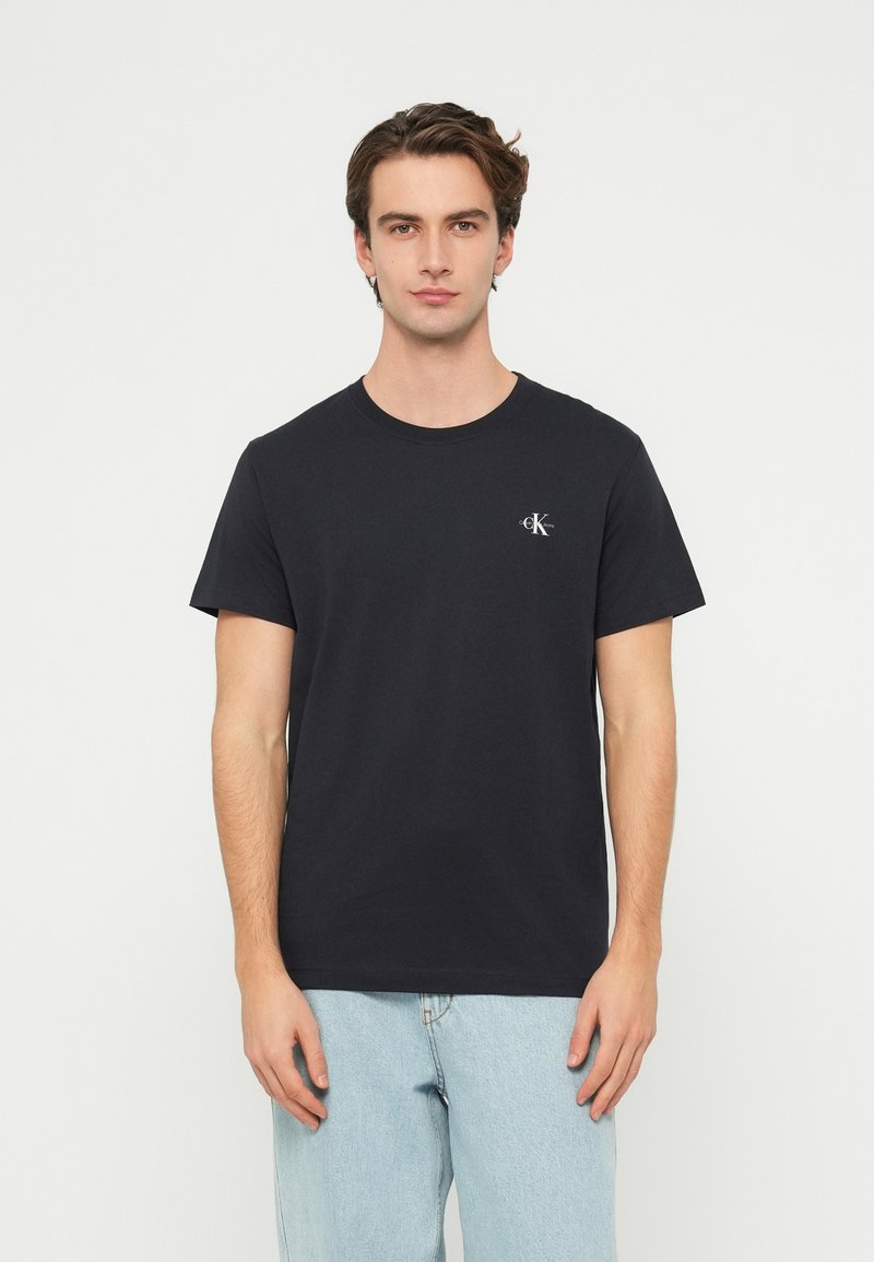 Čierne bavlnené tričko s krátkymi rukávmi, okrúhlym výstrihom a malým logom na hrudi. Hladká textúra, štandardný strih, v kombinácii so svetlomodrými džínsami.