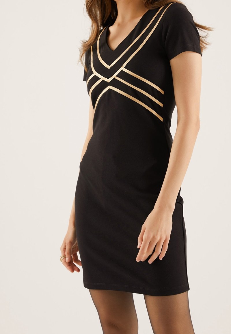 Femme portant une robe noire à manches courtes avec des rayures géométriques beiges au niveau de la poitrine et de la taille haute.