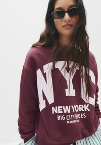 Felpa bordeaux con grande scritta bianca "NYC"; tessuto morbido, vestibilità rilassata, scollo rotondo. Indossata con occhiali da sole neri e un pantalone a righe.