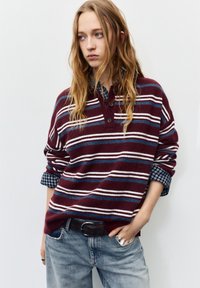 Pulover tricotat cu dungi vișinii, cu dungi albastre și albe, având un guler cu nasturi. Purtat peste o cămașă în carouri albastre și asortat cu blugi deschiși la culoare.