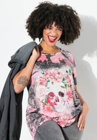 Farverig kortærmet t-shirt med et skull-design og blomsterdetaljer, lavet af blødt stof. Modellen holder en mørkegrå jakke.