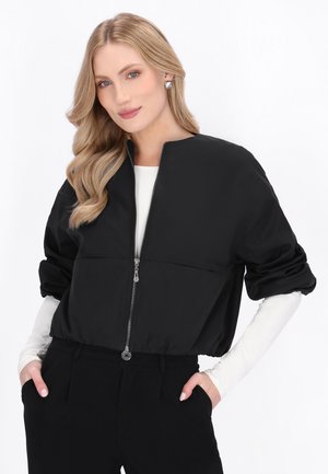 Vrouw met lang blond haar draagt een zwart jack met ritssluiting over een wit shirt met lange mouwen en zwarte broek, poseert met handen in de zakken tegen een witte achtergrond.