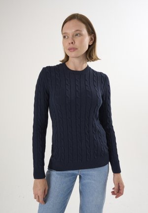 Pull en maille bleu marine présentant des motifs en cable, un col rond et des manches longues. Il est associé à un jean bleu clair.