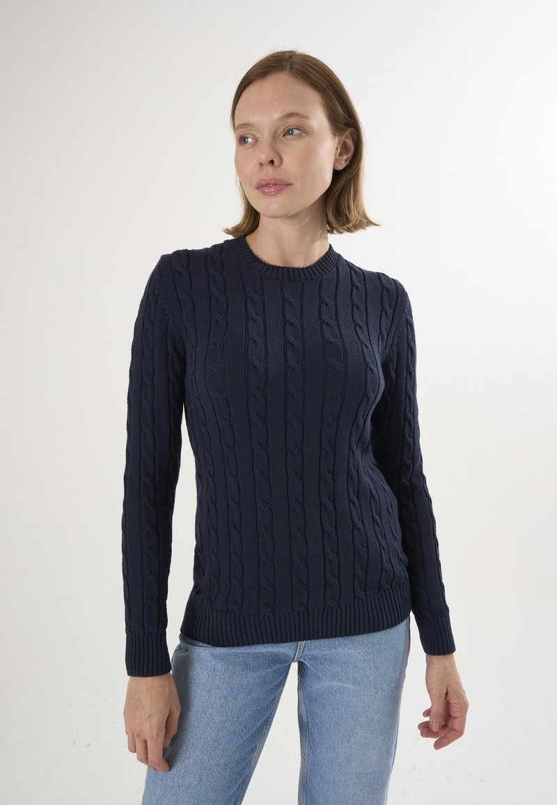 Pull en maille bleu marine présentant des motifs en cable, un col rond et des manches longues. Il est associé à un jean bleu clair.