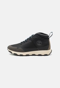 Zapato de senderismo negro impermeable con parte superior de textil, capuchón de goma gris en la punta, cordones azules y suela beige con un patrón de relieve resistente.