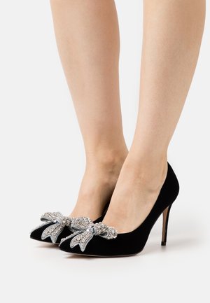 Chaussures à talons hauts en velours noir avec des orteils pointus, ornées de grands nœuds embellis de strass à l'avant. Talon aiguille fin.