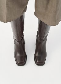 Barna bőr, térdig érő csizmák, négyzetes orral, sima felülettel és oldalsó cipzárokkal, világos háttér előtt.