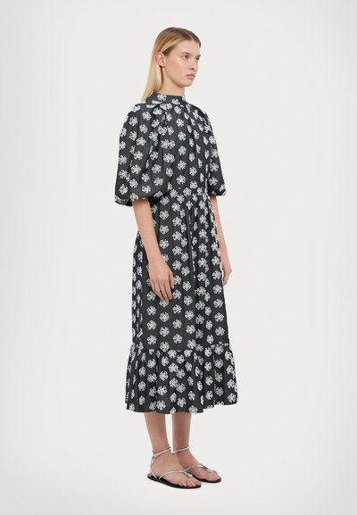 Schwarzes Kleid mit weißem Blumenmuster, kurzen Puffärmeln, hohem Ausschnitt, gestuftem Rüschensaum und leichtem Stoff. Getragen mit silbernen Sandalen.