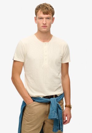 Hellbeige kurzärmliges T-Shirt mit Henley-Ausschnitt und drei Knöpfen; getragen mit beigen Hosen und einem blauen Jeanshemd, das um die Taille gebunden ist.