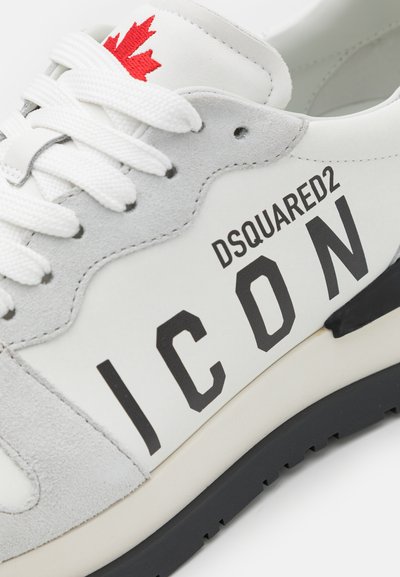 Hvite sneakers med grå semsket skinnoverlegg, svart "ICON"-tekst og rød lønnsløvdetalj. Har flate hvite lissser og en teksturert svart såle.