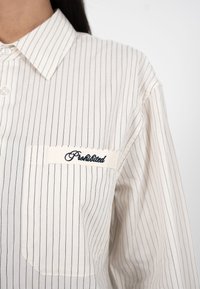 Gros plan sur une chemise blanche à fines rayures avec une poche avant brodée du mot « Interdit » en fil noir cursif.