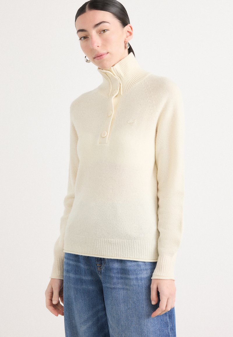 Un pull crème à col montant en tissu doux, doté d'une patte de boutonnage et de poignets côtelés, associé à un jean en denim bleu.