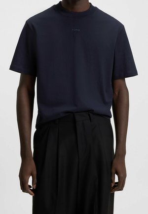T-shirt bleu marine foncé à manches courtes avec petit logo « HUGO » sur la poitrine, porté rentré dans un pantalon noir plissé taille haute.