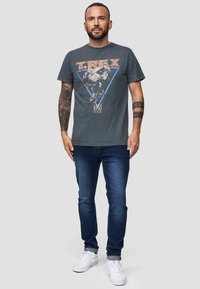 Grijze katoenen t-shirt met een T. Rex-afbeelding binnen een blauwe driehoek, gecombineerd met blauwe spijkerbroek en witte sneakers.
