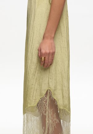 Mano con anillo en espiral dorado descansando junto a un vestido verde claro con un dobladillo de encaje transparente y una textura sutil de puntos.