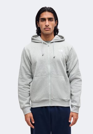 Homme aux cheveux foncés portant un sweat à capuche gris clair zippé avec des poches devant et un pantalon bleu marine, face à l'avant sur un fond uni.