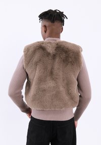 Vest din blană sintetică maro, cu umeri rotunjiti și margine dreaptă, purtat peste un pulover bej cu model ondulat și blugi negri. Fără elemente de prindere vizibile.