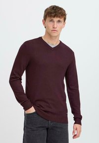 Burgunderfarget V-hals genser laget av mykt strikket stoff, med lange ermer og ribbestrikkede mansjetter, kombinert med mørkegrå jeans.