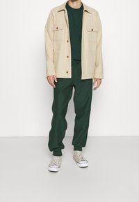 Beige bomulljacka med två bröstfickor, bärs över en mörkgrön t-shirt och matchande gröna byxor; ljusgrå sneakers fullbordar outfiten.