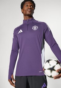 Purple long-sleeve soccer training top with zip neck, white accents, Manchester United logo, and AEROREADY label, paired with a patterned soccer ball.