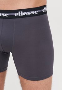 Graue Boxershorts aus elastischem Material mit einem schwarzen Bündchen, auf dem das weiße "ellesse"-Logo prangt. Nahtloses Design und mittellange Beinform bis zum Oberschenkel.