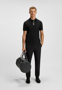 Black polo shirt with short sleeves and a front zipper; dark pants; holding a round black duffel bag with gray straps; smooth textures.
