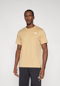 Μπεζ βαμβακερό μπλουζάκι με στρογγυλή λαιμόκοψη, κοντά μανίκια και μικρό λευκό λογότυπο στην αριστερή πλευρά του στήθους. Το μοντέλο φοράει μαύρο παντελόνι.