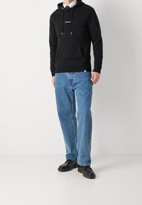 Czarna bluza z kapturem, z kieszenią kangurką, regulowanymi sznurkami i logo z przodu. W połączeniu z jasnoniebieskimi luźnymi dżinsami i czarnymi butami.